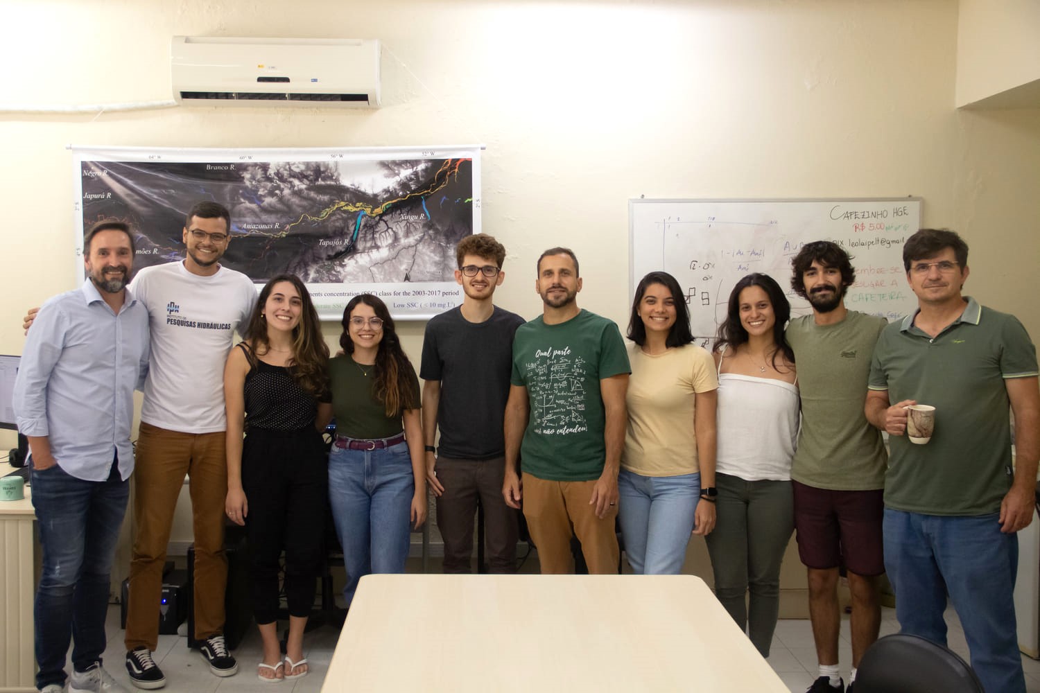 Grupo de dez pessoas aparece posando abraçadas para foto. Todas estão olhando para a câmera, em uma sala com uma mesa no centro e banners na parede.