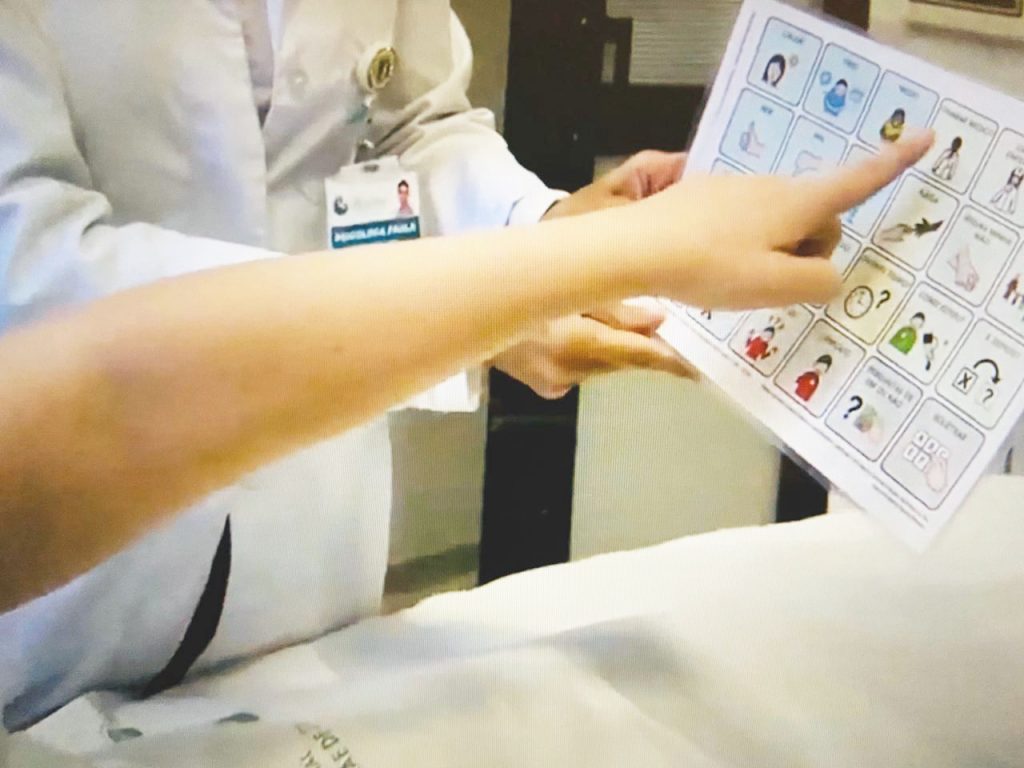 foto colorida de detalhe de uma pessoa que aponta para prancha de comunicação que está nas mãos de outra pessoa com jaleco branco.