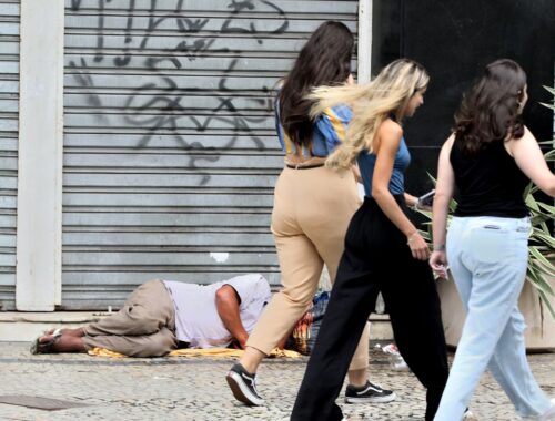 Imagem com morador de rua dormindo sob marquise e pessoas caminhando indiferentes.