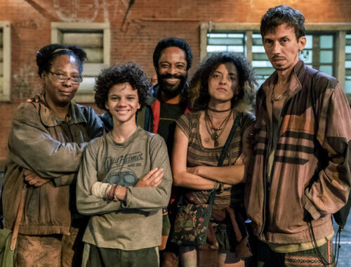 Quatro pessoas de pé, representando personagens empobrecidos. São duas mulheres, uma delas negra, um jovem e dois homens, um deles negro.