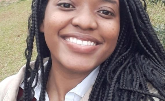 Foto de Melissa sorrindo. Ela é uma mulher negra, de cabelos compridos trançados e está em um lugar ao ar livre, com um gramado e árvores no segundo plano