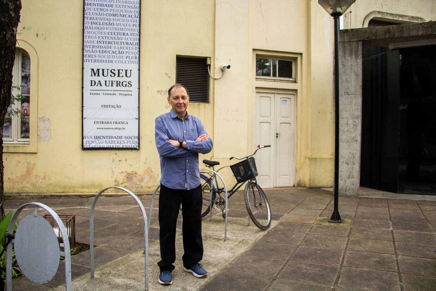 Homem branco, de cabelos grisalhos, usando camisa azul e calça preta, em pé em frente a uma fachada amarela em que se lê Museu da UFRGS, sorrindo levemente e olhando para a câmera