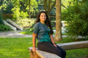 Jovem negra, de pele clara, com olhos verdes e camiseta também verde, com a bandeira do Brasil, posa em jardim com gramado aparente e árvores no fundo