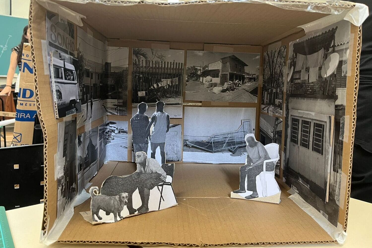 Obra de arte feita com colagens de imagens aparece em primeiro plano. Todas fotos estão dispostas dentro de uma caixa de papelão, mostrando questões do cotidiano, como um senhor fazendo carinho em cachorros ou a imagem da fachada de uma casa.