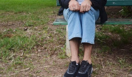 Homem branco, com calça jeans e tênis de corrida aparece sentado em banco de madeira verde. O local que ele está é aberto, com grama no chão e a identidade do homem é preservada, aparecendo somente suas pernas e parte dos braços.