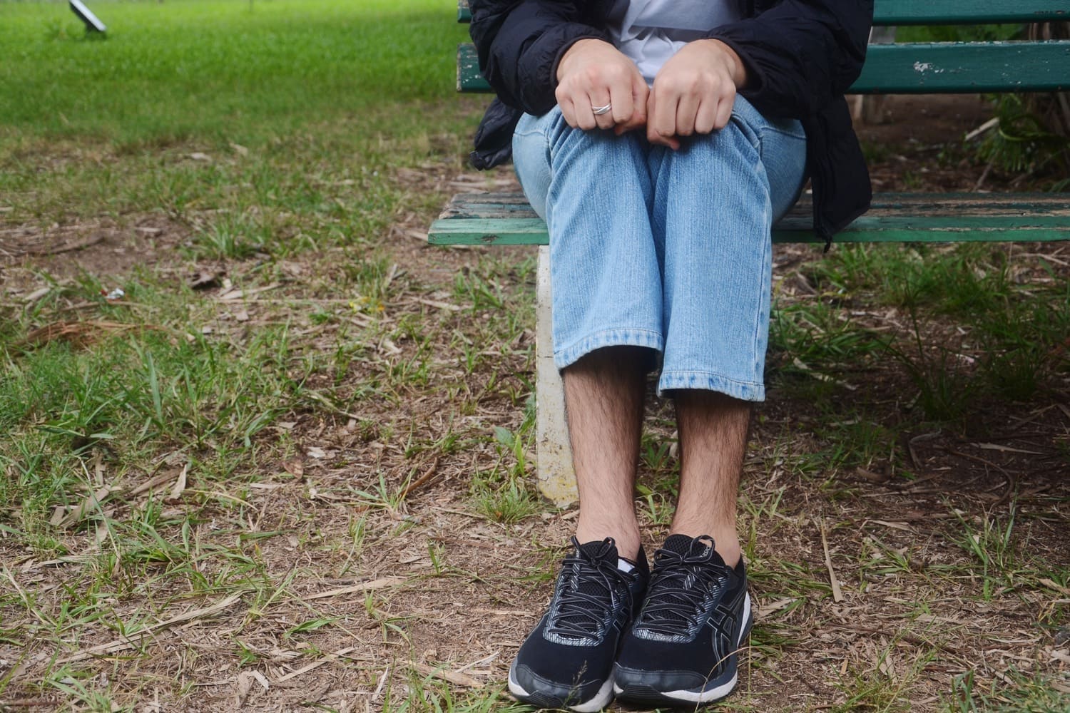 Homem branco, com calça jeans e tênis de corrida aparece sentado em banco de madeira verde. O local que ele está é aberto, com grama no chão e a identidade do homem é preservada, aparecendo somente suas pernas e parte dos braços.