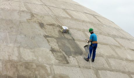 Homem negro, com camiseta e calça azul, capacete verde e luvas aparece de costas enquanto está suspenso por cabos de segurança em um empreendimento de cimento.