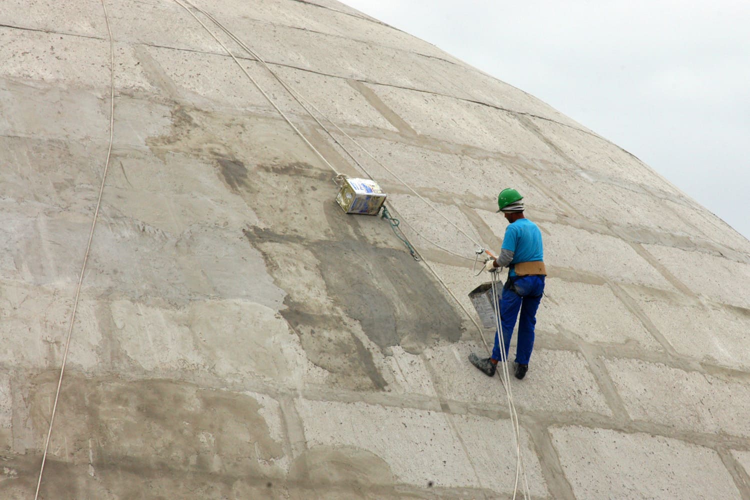 Homem negro, com camiseta e calça azul, capacete verde e luvas aparece de costas enquanto está suspenso por cabos de segurança em um empreendimento de cimento.