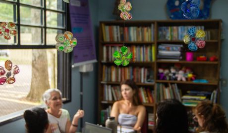 Em primeiro plano flores feitas de garrafa pet são mostradas, penduradas no teto, ao fundo, desfocadas, aparecem 5 mulheres conversando em roda.