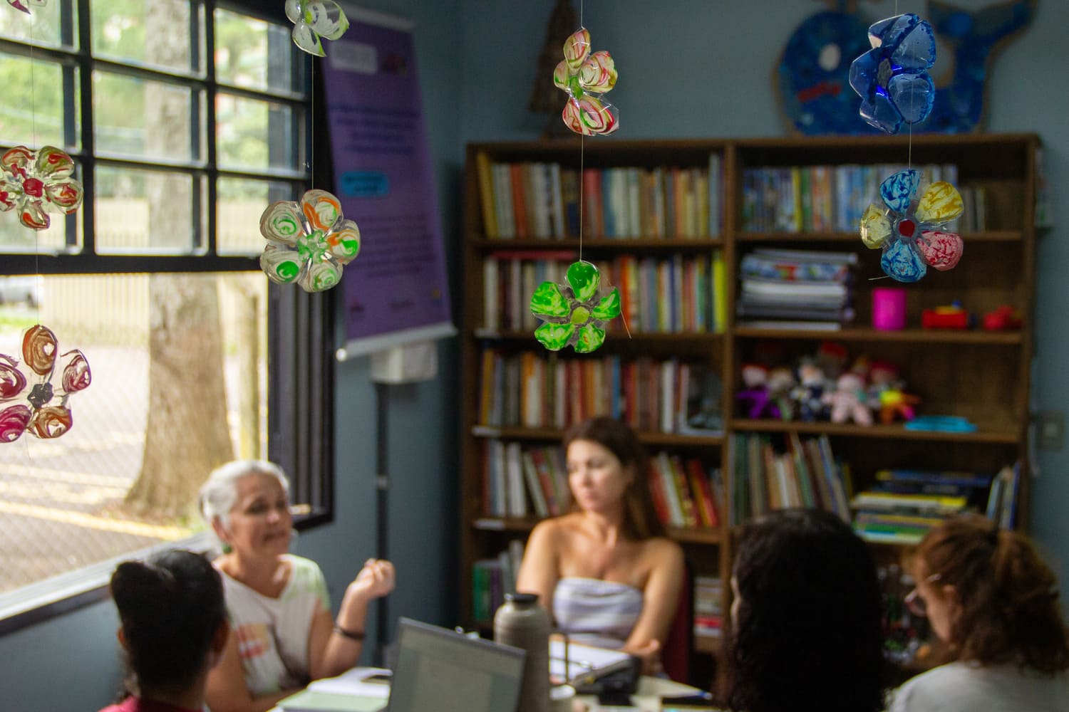 Em primeiro plano flores feitas de garrafa pet são mostradas, penduradas no teto, ao fundo, desfocadas, aparecem 5 mulheres conversando em roda.