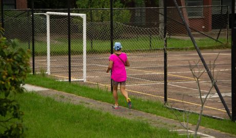 Mulher aparece de costas, enquanto prática corrida, em um ambiente com grama, árvores e também uma quadra de esportes. Ela está de boné, bolsa atravessada no corpo e tênis de corrida.
