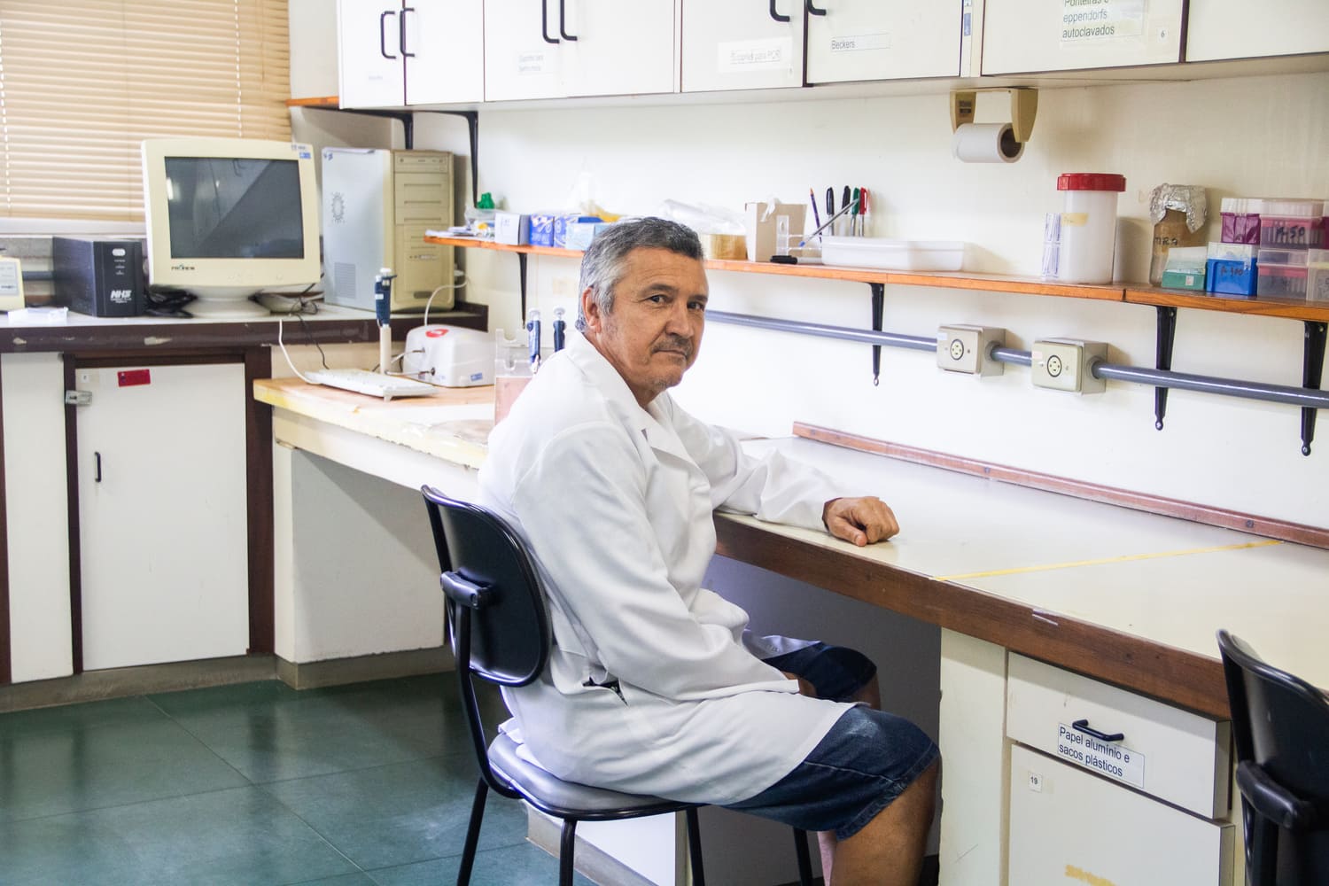 No centro da foto, há um homem branco, de cabelos grisalhos e jaleco branco, sentado em frente a uma bancada de laboratório, olhando diretamente para o fotógrafo.