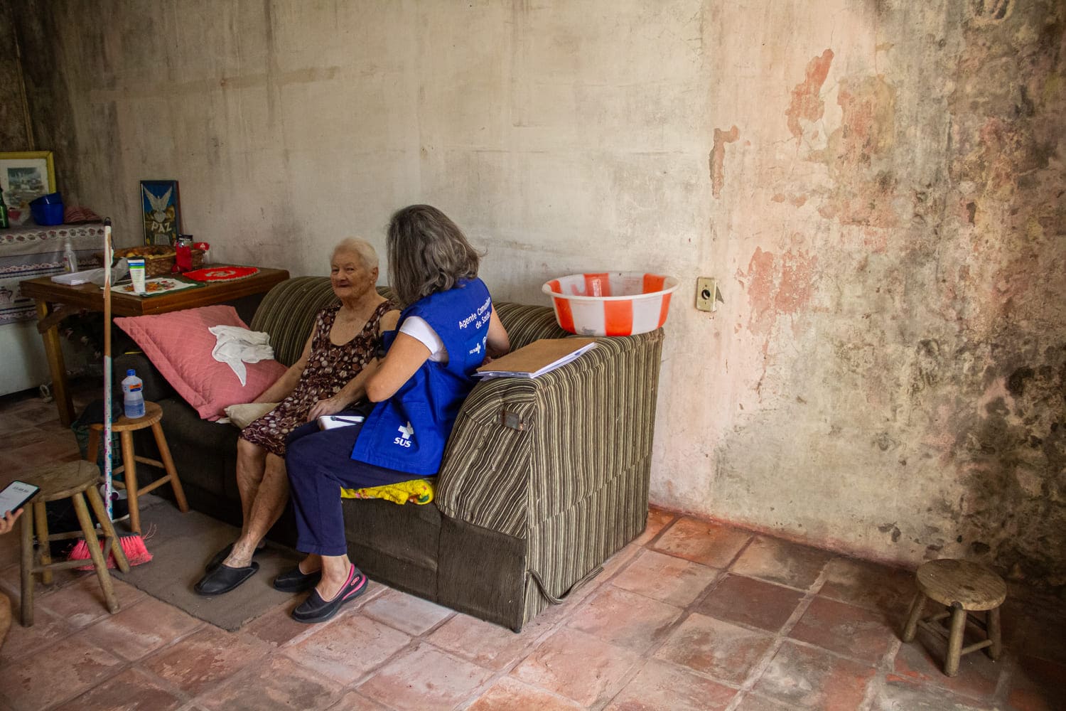Agente comunitária de saúde aparece amparando mulher idosa, enquanto as duas estão sentadas em sofá estampado, cercadas por objetos domiciliares, como bacia de plástico e bancos de madeira.