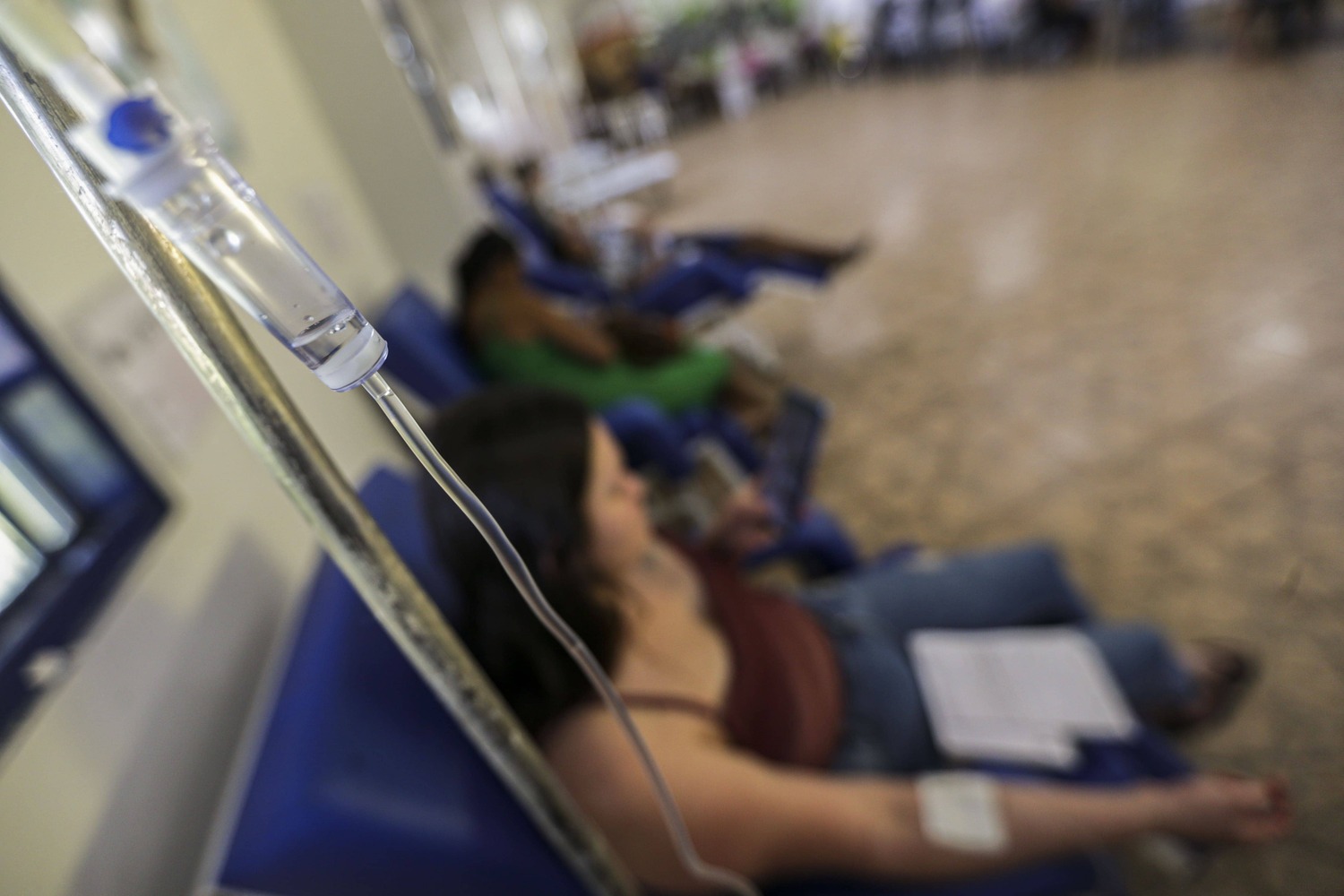 Cerca de três pessoas aparecem sentadas em bancos azuis de um hospital. Elas estão desfocadas, enquanto uma delas, que é uma mulher, com cabelos longos, regata marrom e calça jeans, está com um soro fixado em seu braço. O soro está suspenso em uma barra de ferro, e é o único objeto focado.