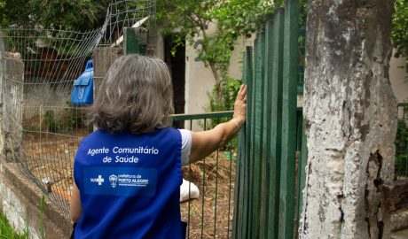 Mulher branca, com cabelos grisalhos e jaleco escrito 'Agente Comunitário de Saúde', aparece de costas, abrindo portão de madeira em uma casa.