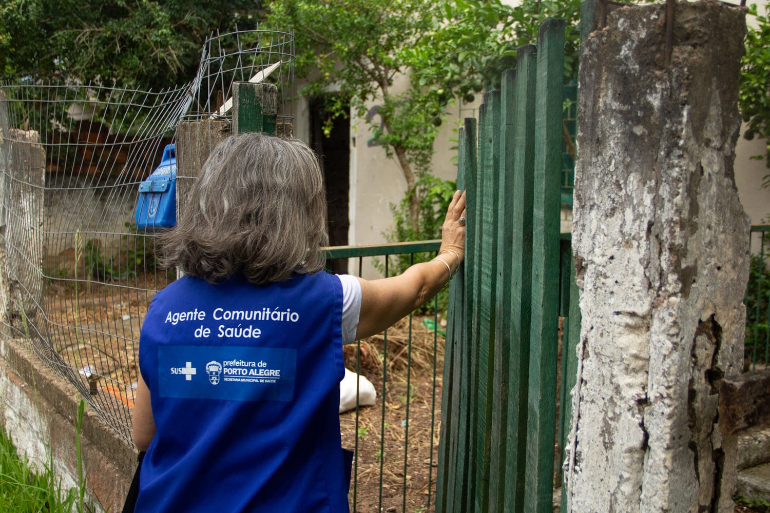 Mulher branca, com cabelos grisalhos e jaleco escrito 'Agente Comunitário de Saúde', aparece de costas, abrindo portão de madeira em uma casa.