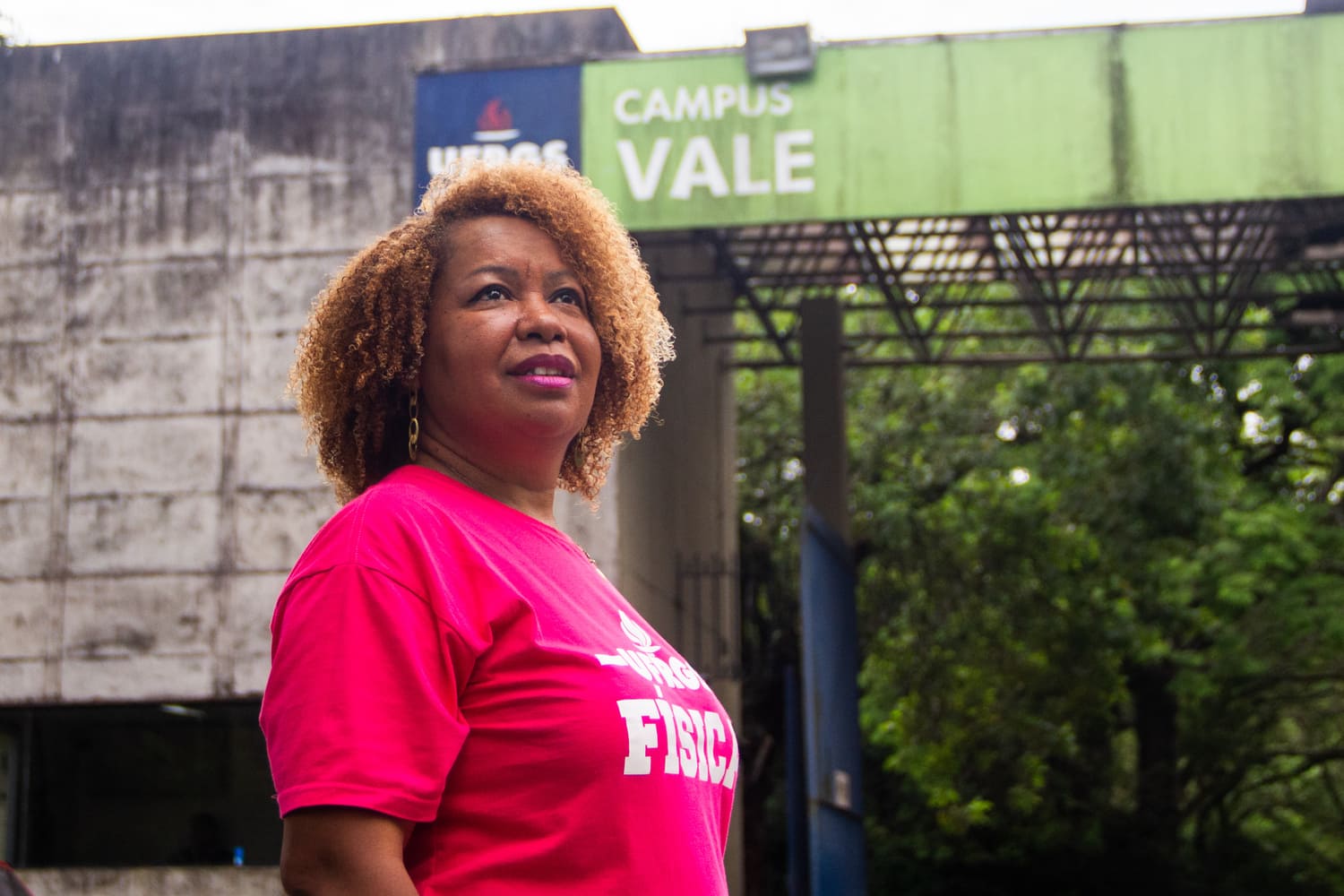 Mulher negra, de cabelos crespos curtos, usando batom e camiseta de cor de rosa, olhando para cima e à direita, em frente a um pórtico que diz UFRGS Campus Vale
