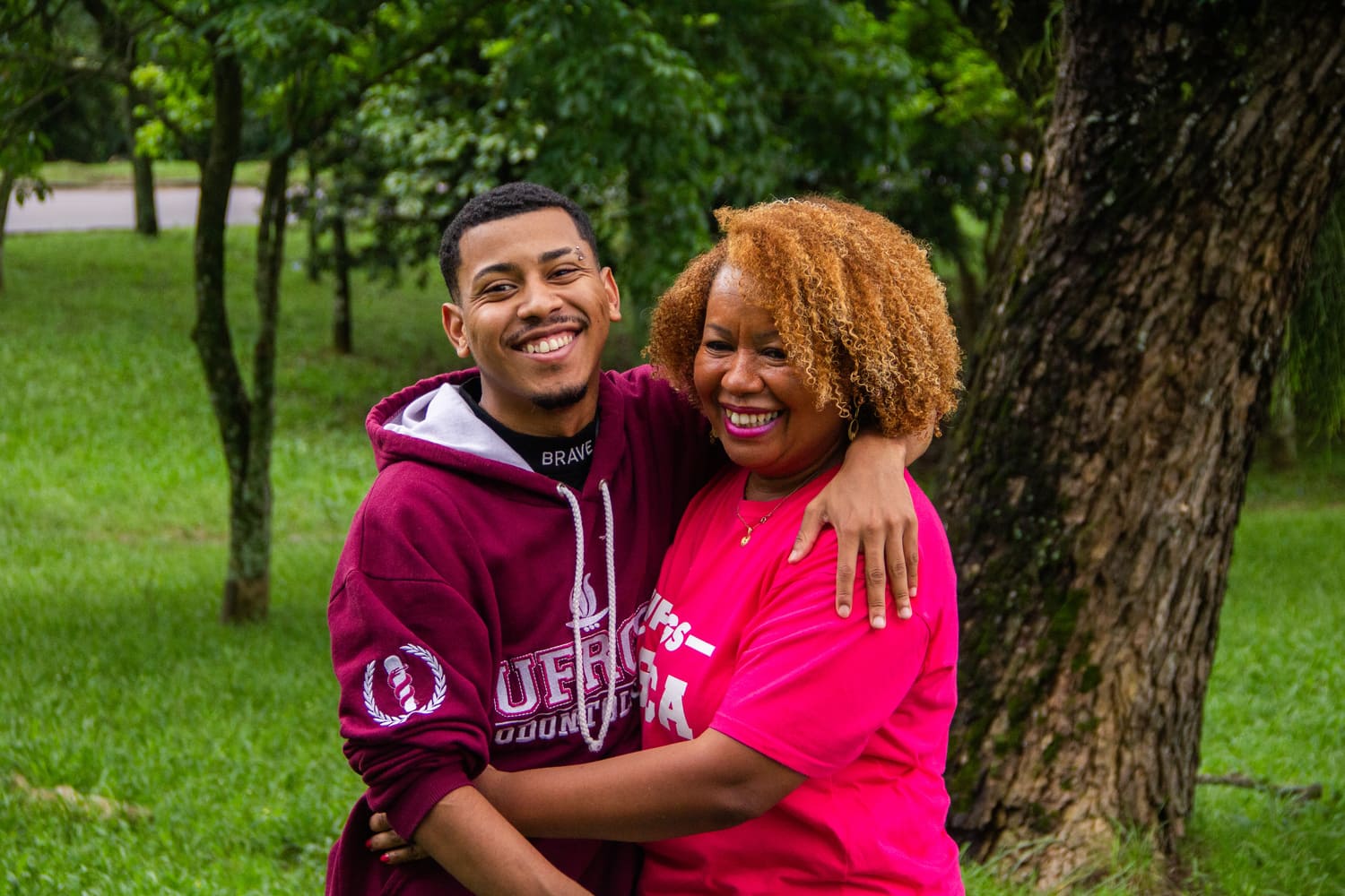À esquerda, rapaz negro, com bigode, cavanhaque e piercing na sobrancelha, usando moletom dizendo UFRGS Odontologia, abraçado em uma mulher negra, de cabelos crespos, usando camiseta cor de rosa. Ambos sorriem para a foto. Ao fundo, vegetação com árvores e grama.