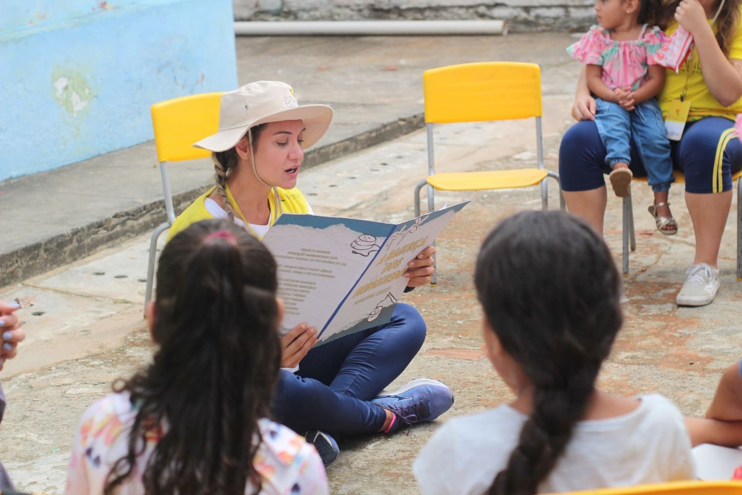 Mulher branca, loira e com uma trança no cabelo, lê livro para grupo de crianças, enquanto está sentada de pernas cruzadas no chão. À sua volta estão dispostas cadeiras amarelas, que estão sendo utilizadas para os ouvintes sentarem.