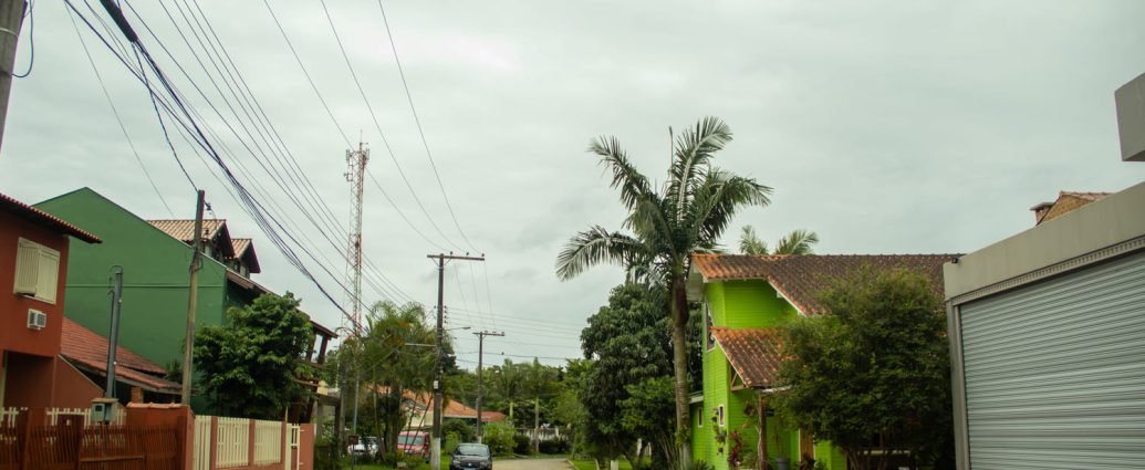 Rua de pedras aparece em primeiro plano, com casas nas duas extremidades. Palmeiras e outras árvores compõem o local, junto a carros e fios de eletricidade.