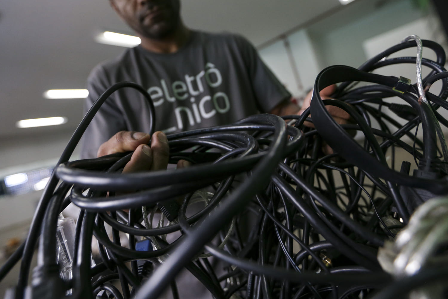 Homem negro, usando camiseta com a palavra "Eletrônico" estampada, segura vários fios de diferentes eletrônicos. O indivíduo está desfocado e seu rosto não aparece, dando destaque aos fios e a suas mãos.