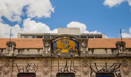 Em primeiro plano, prédio do Museu da Universidade Federal do Rio Grande do Sul, localizado no centro de Porto Alegre. Ele é bege, com detalhes coloridos. No fundo, aparece o céu azul e também o topo do prédio da Faculdade de Educação.