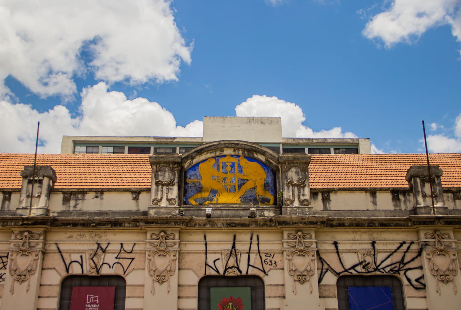 Em primeiro plano, prédio do Museu da Universidade Federal do Rio Grande do Sul, localizado no centro de Porto Alegre. Ele é bege, com detalhes coloridos. No fundo, aparece o céu azul e também o topo do prédio da Faculdade de Educação.