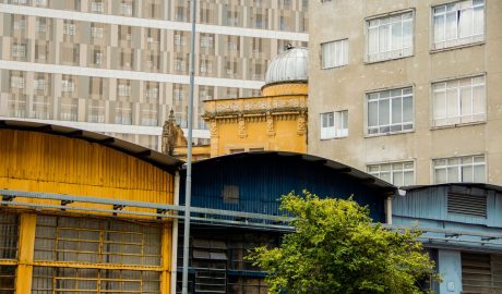 Armazéns nas cores amarelo e azul aparecem em primeiro plano, junto a árvore. Já no fundo, está prédio amarelo, com cúpula na parte de cima.