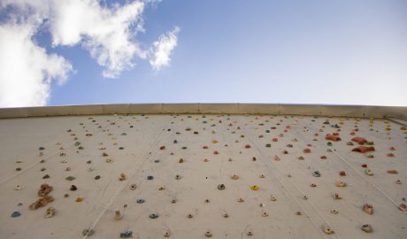 Prédio principal do Campus da Escola de Educação Física, Fisioterapia e Dança, onde há uma parede de escalada, com apoios para as mãos nas cores amarelo, azul, verde e vermelho. Além do prédio bege, o céu azul aparece na imagem, que foi tirada de baixo para cima.