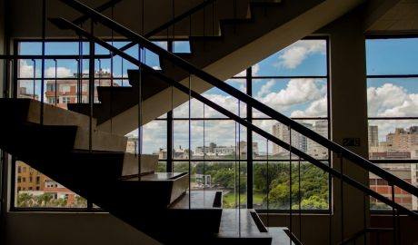 Em primeiro e segundo plano, degraus da escada da Faculdade de Educação da UFRGS. No fundo, aparece a janela do local, que se localiza no alto do nono andar, com paisagem ensolarada.