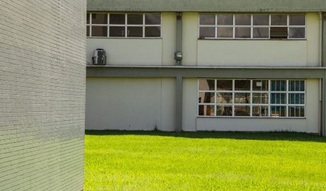 No fundo, Ginásio da Faculdade da Escola de Educação Física, Fisioterapia e Dança, onde janelas aparecem em destaque, com prédio em tons de cinza. Já em primeiro plano, gramado verde e parede de outro prédio, com ladrilhos cinzas. Tudo em dia ensolarado.