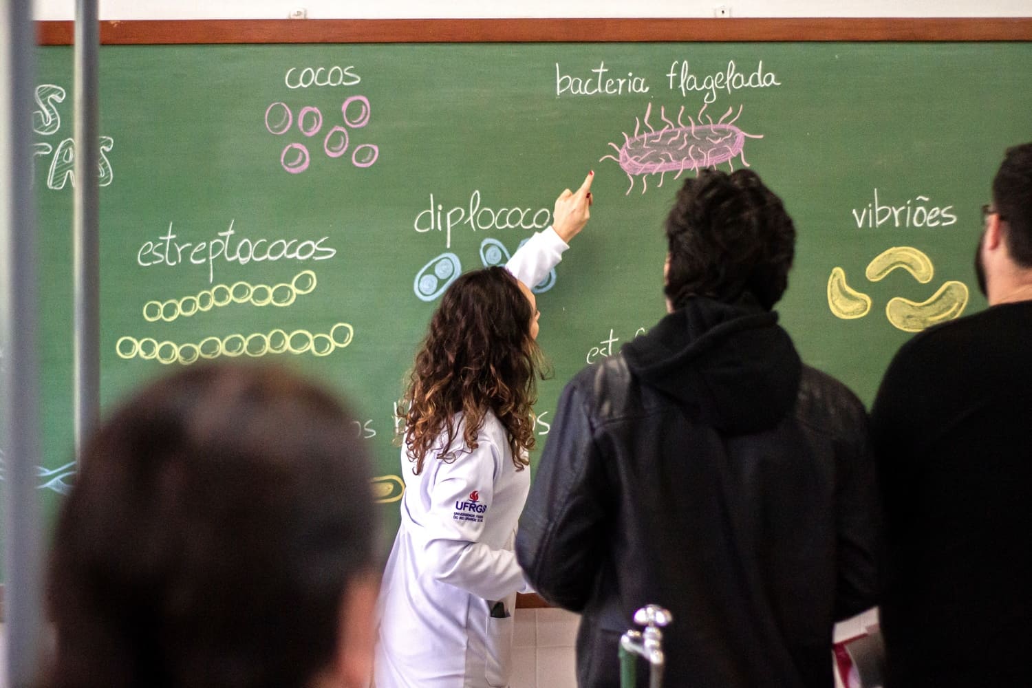 Imagem mostra professora com cabelos loiros e jaleco branco apontando o dedo para informação que está escrita em quadro de sala de aula. Na informação diz: "bactéria flagelada", com desenho ilustrando a mesma abaixo. E em volta da professora, estão alunos acompanhando os ensinamentos.