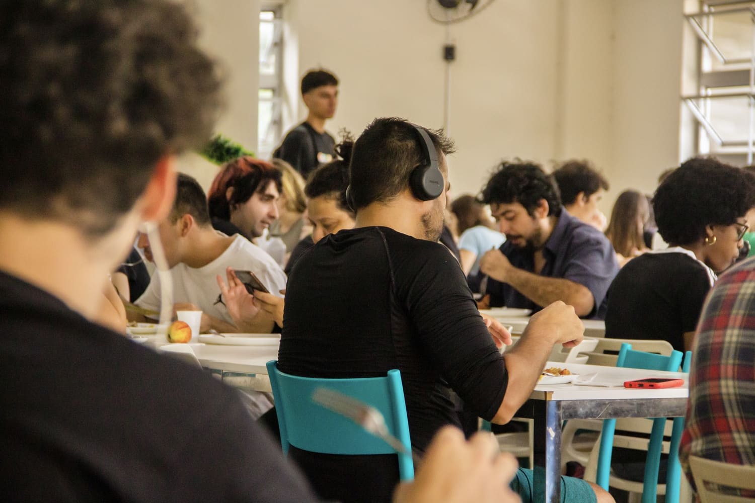 Foto mostra em destaque estudante com fones de ouvido e camiseta, ambos na cor preta, sentado em mesa branca, com cadeiras azuis, comendo durante o almoço. Em sua volta, estão outros estudantes fazendo o mesmo. O ambiente tem paredes brancas e janelas ao fundo.