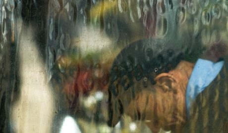 Homem com cabeça baixa, junto a uma janela molhada de chuva, solidão.