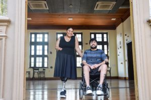 Na fotografia aparecem duas pessoas, uma mulher à esquerda, com pernas mecânicas e deficiência em um braço; à direita, um homem cadeirante. Os dois são fontes para reportagem sobre acessibilidade para estudantes com deficiência (PCDs) na UFRGS