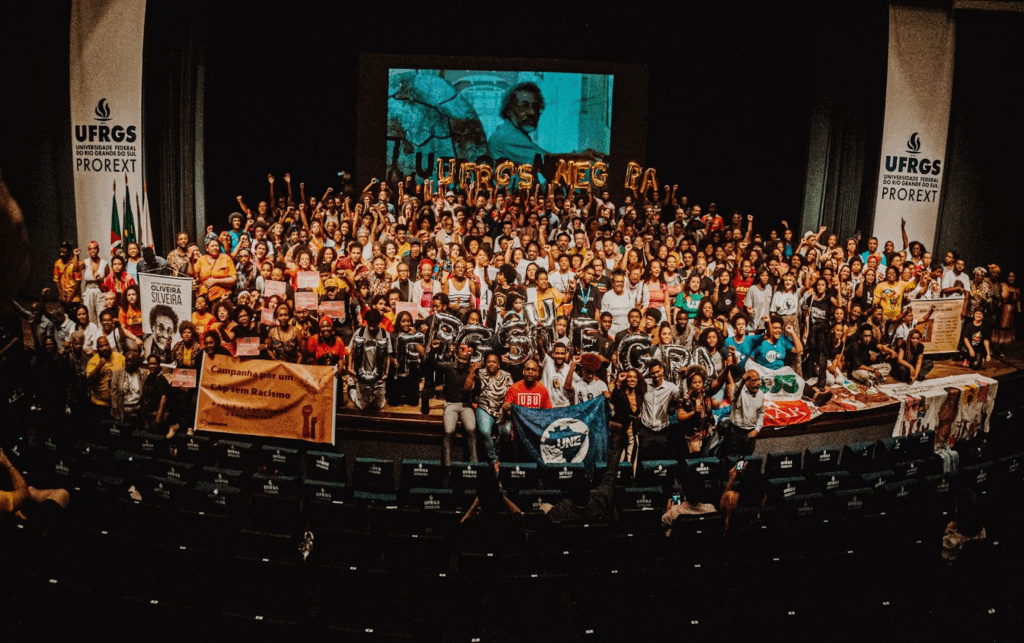 Conjunto de pessoas negras em cima do palco do Salão de Atos da UFRGS com a foto do Oliveira Silveira ao fundo, balões compondo a palavra UFRGS NEGRA, no evento Novembro Negro UFRGS.