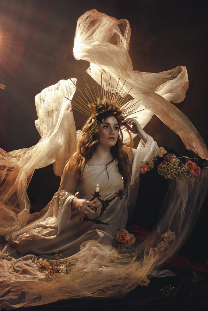 Fotografia de uma mulher sentada no chão segurando um candelabro, enquanto está apoiada com o braço em um pequeno pilar, com flores em cima. Ela usa uma coroa com flores e espetos pequenos.