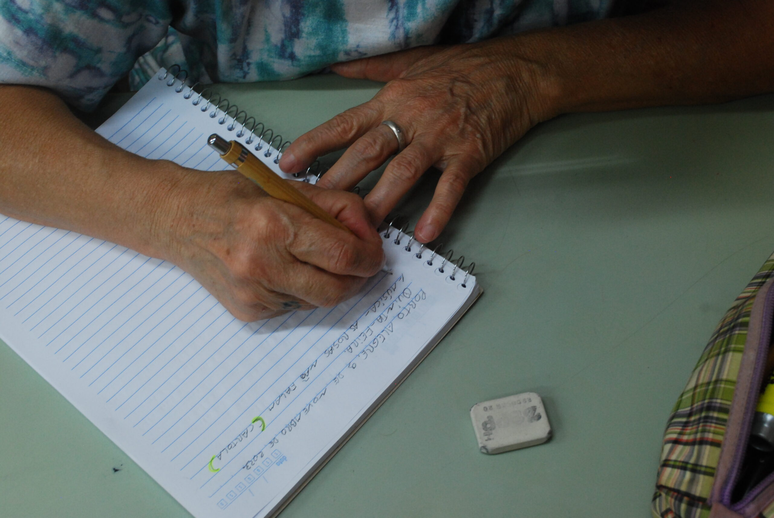 Mãos de Ilce segurando lapiseira e escrevendo em um caderno.