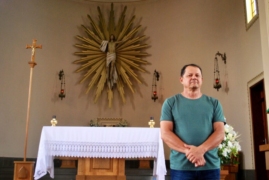 Homem de 56 anos dentro de igreja, com as mãos em frente ao corpo e escultura de Jesus Cristo ressuscitado atrás dele