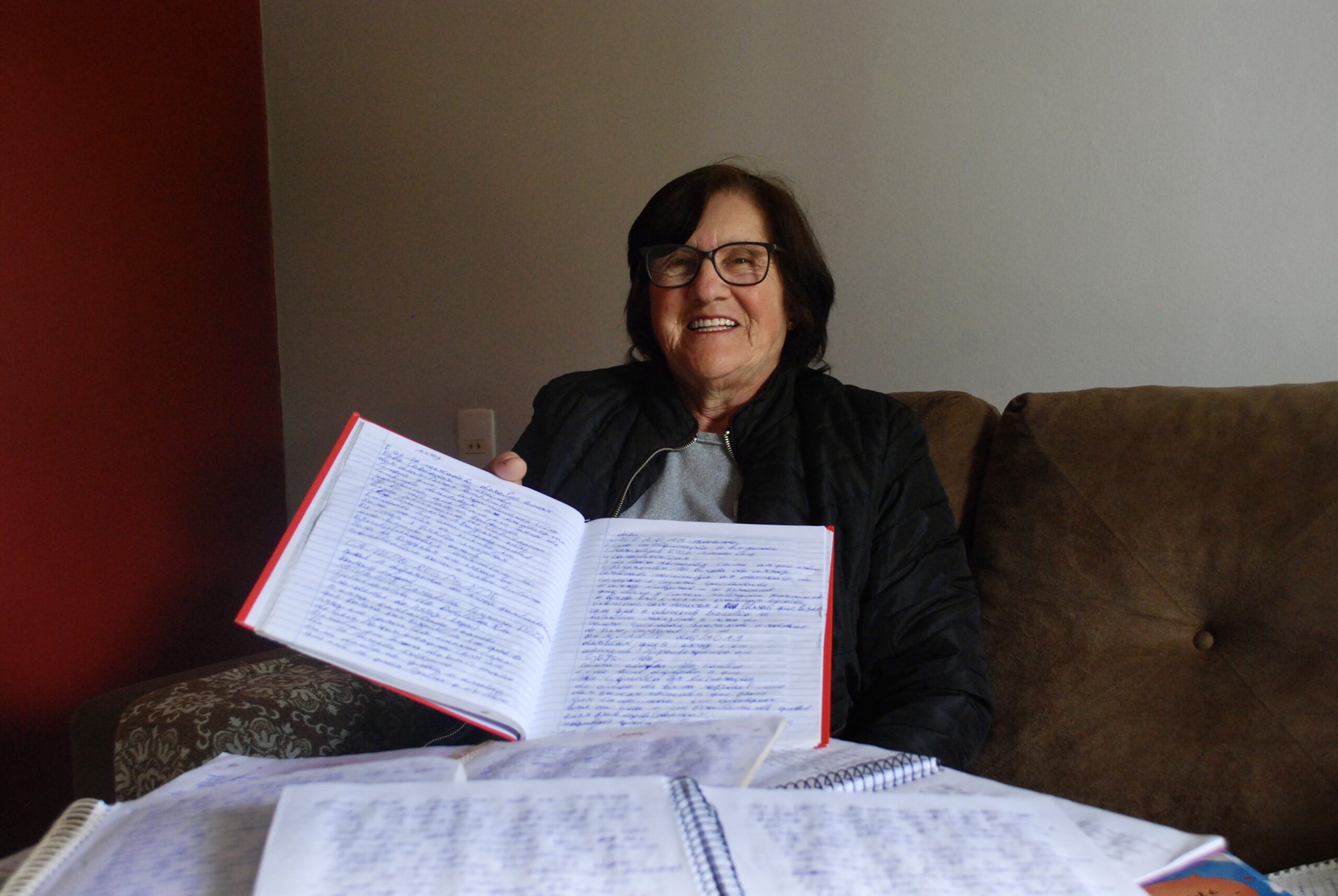 Sueli, sentada em um sofá, segurando os cadernos que utilizava para transcrever textos.