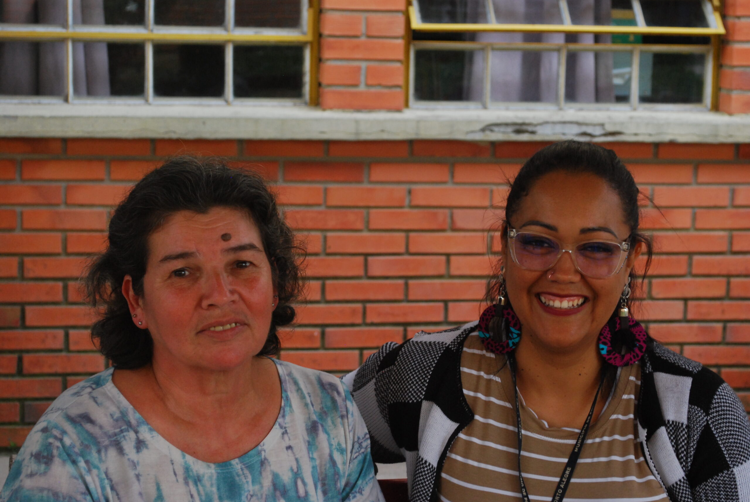 Estudante Ilce e professora Perla sentadas, lado a lado, em banco colorido da escola.