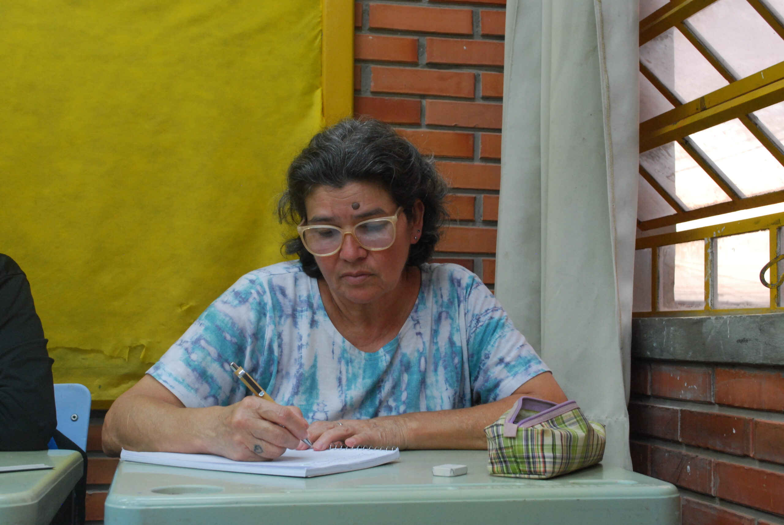Ilce escrevendo no caderno, sentada em uma classe.
