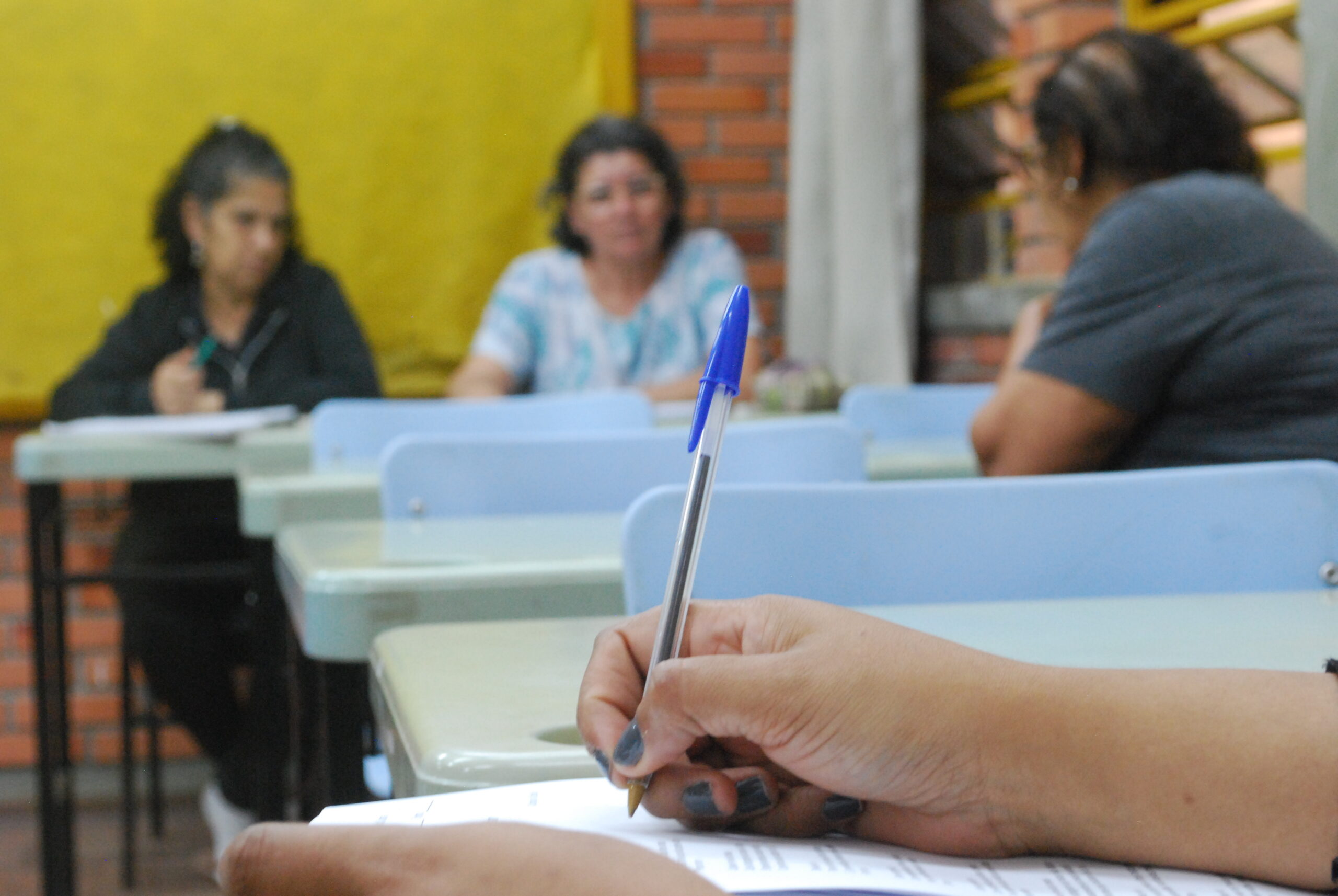 Em foco, mão segurando caneta. Ao fundo, alunas conversando em sala de aula.