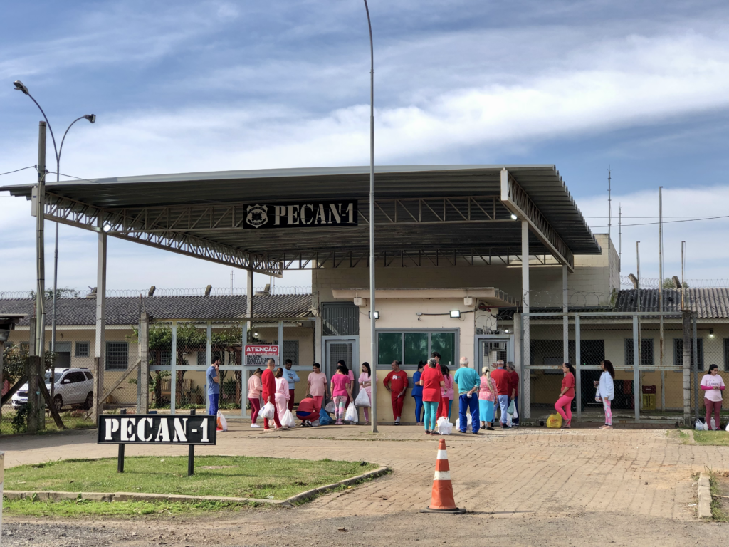 Movimentação em frente ao prédio um do Presídio Municipal de Canoas, com filas de pessoas indo visitar pessoas privadas de liberdade. O prédio tem apenas um andar, paredes bege e teto cinza. Em volta da entrada, há placas de aviso como "atenção" e "cuidado", cones e câmeras de segurança. Fim da descrição.