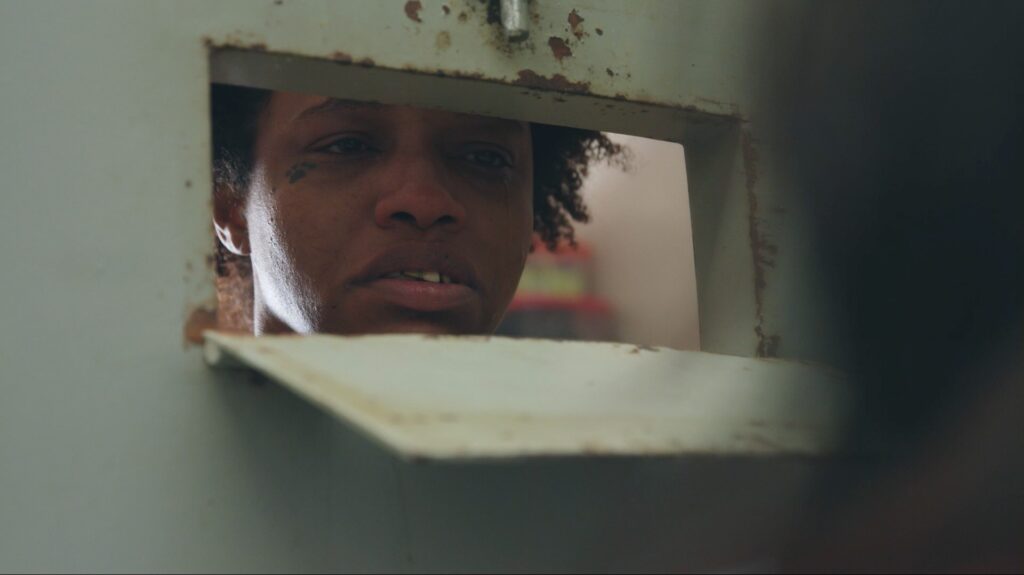 Pôster do documentário "Olha para Elas", de Renato Dornelles e Tatiana Sager. Na foto, uma mulher privada de liberdade olha para a câmera por meio de uma portinhola. Fim da descrição.