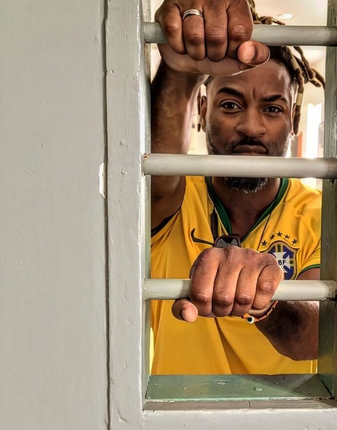 Imagem de Rodrigo Sabiah usando uma camisa da Seleção Brasileira de Futebol, ele olha para a câmera por meio da abertura de uma porta de uma unidade prisional enquanto segura as grades dessa abertura, possui um semblante sério e confiante, demonstrando a sua determinação à luta pela ressocialização dos egressos. Fim da descrição.